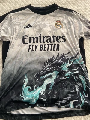 Real Madrid draktröja Mbappé #10 - Unik Real Madrid matchtröja med drakmotiv och Mbappé #10 på ryggen. Tröjan är svart, grå och turkos med klubbmärke och sponsortryck framtill. Tillverkad i lätt och ventilerande material, perfekt för fotbollsträning eller support.