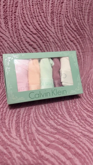Calvin Klein trosor i pastellfärger - Fempack trosor från Calvin Klein i mjuka pastellfärger: ljusrosa, persika, mintgrön, lila och vit. Ribbad bomull med bred resår i midjan och diskret Calvin Klein-logga. Söt spetskant runt benen för extra detalj. Perfekt för dig som gillar bekväma och snygga underkläder.
