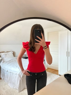 Röd volang topp med v-ringning - Snygg röd topp med djup v-ringning och volangdetaljer vid axlarna. Modellen är ärmlös och har en croppad passform som sitter tight. Tillverkad i mjuk bomull, perfekt för att lyfta vilken outfit som helst.