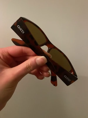 Gucci bruna solglasögon i retrostil - Snygga solglasögon från Gucci med brunmelerat, fyrkantigt båge och mörka glas. Klassisk logga på båda skalmarna. Perfekt accessoar för dig som vill sticka ut med en exklusiv och trendig look.