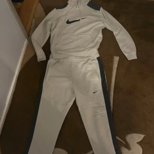 Nike tracksuit - Nike hoodie i vit med svarta axelpartier och stor svart logga på bröstet. Tröjan har huva med snörning, magficka och långa ärmar. Materialet är mjuk bomull och polyester, perfekt för chill eller träning. Snygg och sportig look med normal passform. Ett litet sår innaför byxorna som ej syns utanför