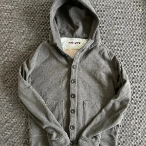 Grå cardigan med knappar från NN07 - Snygg grå hoodie från NN07 med knappar hela vägen framtill och två stora fickor. Tröjan har huva och är långärmad, perfekt för chill dagar. Mjuk bomullskänsla och stilren design som funkar till jeans eller joggers.