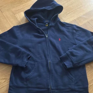 Marinblå hoodie från Polo Ralph Lauren - Snygg marinblå hoodie från Polo Ralph Lauren med röd broderad logga på bröstet. Tröjan har huva, dragkedja framtill och två fickor. Mjuk och skön bomullskvalitet, perfekt för chill dagar. Klassisk stil med lång ärm och ribbade muddar.