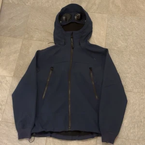 CP Company jacka - Säljer en mörkblå fleecejacka från C.P. Company med ikoniska goggles i huvan. Jackan har två snedställda fickor med dragkedja, hel dragkedja framtill och en stilren, sportig look. Jackan är av fleece material på insidan och jackan, normal passform kan till och med passa S!