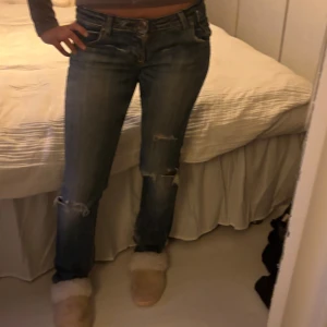 blå jeans med slitningar - Säljer ett par blå bootcut jeans med snygga slitningar på knäna och klassisk femficksdesign. Jeansen har en låg midja, tillverkade i jeansmaterial med lätt tvättad look. Perfekt för dig som gillar en avslappnad och cool stil.