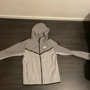 Grå Nike vindjacka med huva - 🔥 Grå Nike Tech Fleece / vindjacka – clean & sportig! Snygg grå Nike-jacka med svart dragkedja, huva och stilrena detaljer. Logga på bröstet och fickor med dragkedja på sidorna. Perfekt för dig som gillar en sportig look och vill ha något lätt att slänga på sig 👟  💰 600 kr – snabb affär! 