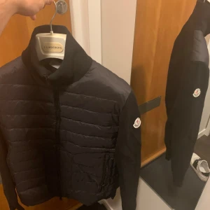 Cardigan från Moncler - Svart pufferjacka från Moncler med quiltad front och stickade ärmar. Jackan har hög krage, dragkedja och Moncler-logga på ärmen. Materialmix av polyester och bomull ger en skön och modern känsla. Perfekt för dig som gillar stilrena och exklusiva plagg.