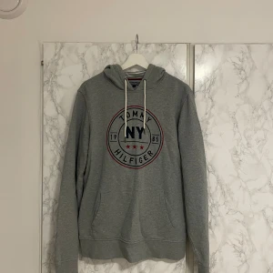 Grå hoodie Tommy Hilfiger NY M - Grå hoodie från Tommy Hilfiger med broderad NY-logga och röda stjärnor på bröstet. Tröjan har vit snörning i huvan, känguruficka och randigt foder i huvan. Perfekt för dig som gillar streetstyle och klassisk design.