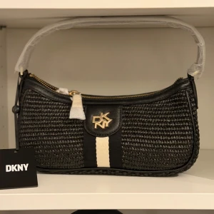Svart baguetteväska från DKNY - Snygg svart baguetteväska från DKNY med flätad struktur och guldfärgad logga framtill. Väskan har en vit och svart randig detalj på framsidan, dragkedja i guld och är tillverkad av polypropylen, polyester och skinn. Perfekt för dig som gillar stilrena accessoarer.
