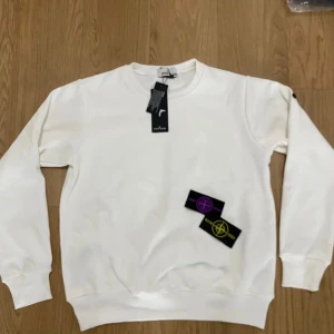 Vit sweatshirt från Stone Island - Säljer en clean vit sweatshirt från Stone Island med klassisk rund hals och ribbade muddar. Tröjan kommer med två utbytbara logopatchar i lila och gult. Perfekt för dig som gillar stilrena och ikoniska streetwear-plagg.