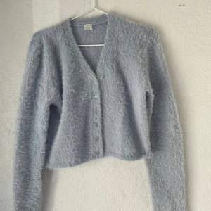 Ljusgrå fluffig kofta från Lindex - Supermjuk och fluffig ljusgrå kofta från Lindex med v-ringning och knappar framtill. Koftan har lång ärm och är tillverkad i ett stickat material som känns riktigt cozy. Perfekt att slänga på sig när det är kyligt ute.