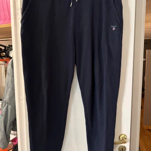 Mörkblå mjukisbyxor från Gant strl: 2XL - Snygga mörkblå mjukisbyxor från Gant med broderad logga på benet och guldfärgade detaljer på snörningen. Byxorna har ribbade muddar vid bensluten och är gjorda i mjukt bomullsmaterial. Perfekta för chill. Mindre noppor kan förekomma.