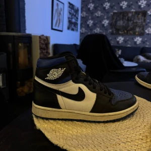 Nike Air Jordan 1 svart/blå sneakers - Säljer ett par Nike Air Jordan 1 sneakers i svart, blått och vitt. Skorna har högt skaft, klassisk snörning och rund tå. Ikonisk Air Jordan-logga på sidan och svart Swoosh. Tillverkade i skinn med vit mellansula och blå yttersula. Perfekt för dig som gillar streetwear och sneakers.