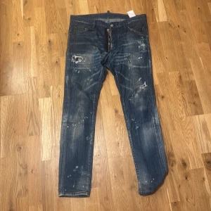 Dsquared2 Cool Guy jeans blå - Snygga Dsquared2 Cool Guy jeans i blå tvätt med slitningar och färgstänk för en rå streetlook. Klassisk femficksmodell, raka ben och låg midja. Tillverkade i Italien av bomull. Perfekta för dig som gillar edgy och trendig stil.