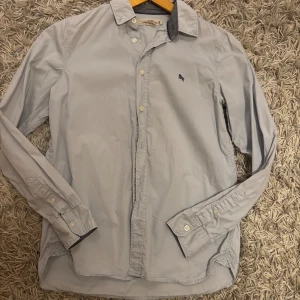 Ljusgrå skjorta från L.O.G.G. - Snygg ljusgrå skjorta från L.O.G.G. med klassisk krage och knappar framtill. Skjortan har lång ärm och ett mörkblått broderat lejon på bröstet. Tillverkad i mjuk bomull som känns skön mot huden. Perfekt för dig som gillar stilrena och enkla plagg.