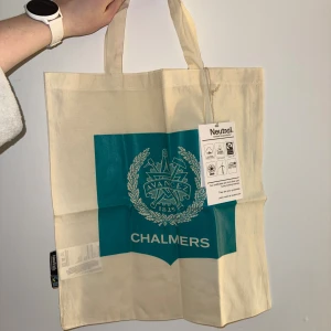 Chalmers shoppingväska - Snygg shoppingväska i beige bomull med turkos tryck och Chalmers emblem. Väskan har två handtag och är perfekt för att bära böcker eller mat. Tillverkad av certifierad ekologisk bomull och Fairtrade-märkt. Enkel och stilren design med tydlig Chalmers-logga.