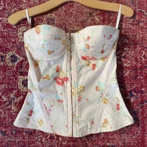 Blommig korsett med axelband - Säljer en ljus korsett med blommigt mönster i rött, gult och grönt. Toppen har formade kupor, smala axelband och markerad midja. Perfekt för dig som gillar romantisk och feminin stil. Stängning framtill och lätt utsvängd nederkant.