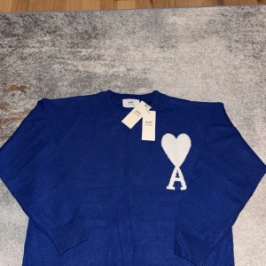 Blå stickad tröja från Ami Paris - Blå stickad tröja från Ami Paris med rund hals och ribbade muddar. På bröstet finns ett stort vitt hjärta med bokstaven A, vilket ger en unik och trendig look. Perfekt för dig som gillar stilrena detaljer och vill sticka ut.