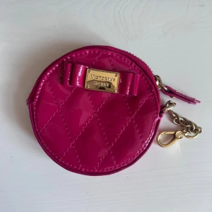 Rosa quiltad plånbok Victoria's Secret - Snygg rund plånbok från Victoria's Secret i glansig rosa färg med quiltat mönster och en söt rosett framtill. Metallplakett med logga och guldfärgad kedja med karbinhake. Perfekt för småsaker och nycklar.