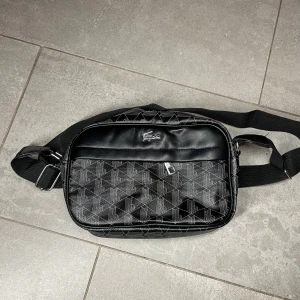 Svart axelväska från Lacoste - Snygg svart axelväska från Lacoste med geometriskt mönster i vitt. Väskan har justerbar rem, dragkedja och en metall-logga framtill. Materialet är skinnimitation och textil, perfekt för dig som vill ha en stilren och praktisk väska med cool design.