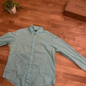 Ljusblå skjorta från Ralph Lauren XL - Snygg ljusblå skjorta från Ralph Lauren i storlek XL. Klassisk design med knappar framtill och lång ärm. Ikonisk rosa broderad logga på bröstet. Skjortan är gjord i mjuk bomull och har en stilren krage. Perfekt för dig som gillar preppy stil och vill ha något fräscht i garderoben.