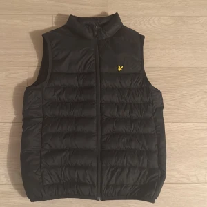 Svart dunväst från Lyle & Scott - Svart dunväst från Lyle & Scott med gul broderad fågel på bröstet. Västen har quiltad design, hög krage och dragkedja framtill. Perfekt att slänga över en hoodie för extra värme. Materialet är polyester och dun, vilket gör den både lätt och varm.