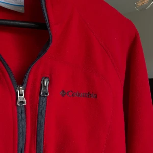 Röd fleecejacka från Columbia - Riktigt snygg röd fleecejacka från Columbia med svart dragkedja och bröstficka. Jackan har hög krage och lång ärm, perfekt för kyliga dagar. Diskret logga broderad på bröstet. Enkel och sportig look som funkar till många stilar.