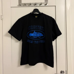 Svart Cortez t-shirt med blått tryck - Svart t-shirt från Cortez med stort blått tryck framtill där det står 'Cortez Rules The World' och en bild av ett skepp. Klassisk passform med rund hals och korta ärmar. Perfekt för dig som gillar streetwear och vill sticka ut med en cool printad tee.