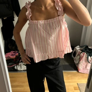 Rosa och vit randig linnetopp med volang - Supersöt linnetopp i vitt med rosa ränder och volangdetaljer längs axelbanden och nederkanten. Toppen har en luftig och loose passform med breda axelband och rynkade detaljer. Perfekt för varma dagar och en chill look.