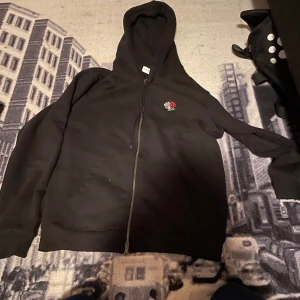 Svart hoodie från Les Deux med ros - Svart hoodie från Les Deux med dragkedja och huva. På bröstet finns ett broderat motiv med en vit och en röd ros. Tröjan har långa ärmar, snörning vid huvan och en stor ficka framtill. Perfekt för chill dagar och streetwear vibes.