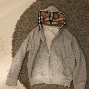 Grå Burberry hoodie med rutig huva - Säljer en grå hoodie från Burberry i storlek M men skulle säga att den är mer storlek S Tröjan har dragkedja framtill, snörning vid huvan och den klassiska Burberry-rutiga insidan på huvan. Mjuk bomullskänsla och två fickor framtill. Perfekt för dig som gillar stilrena och exklusiva detaljer. Hör av er vid frågor eller annat