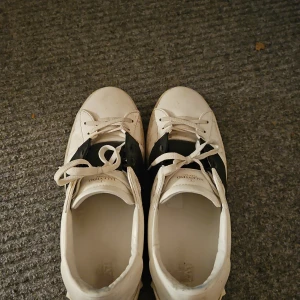 Valentino Open Garavani sneakers (äkthetsbevis) - Säljer ett par Valentino Open Garavani sneakers i vitt och svart skinn med ikoniska nitar runt hälen. Skorna har snörning och platt sula, med en chunky och modern siluett. Medföljer originalkartong, dustbags och extra skosnören. Skorna är i bra skick men lite slitage vid tån på höger sko. Skorna är smutsiga men går att tvätta bort enkelt med ljummet vatten. Pris kan diskuteras! Hör av er om intresserad!