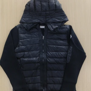 Svart Moncler cardigan  - Svart  Moncler med quiltad framsida och huva. Ärmarna är stickade och har en liten ficka med dragkedja på vänster arm. Jackan har hel dragkedja framtill och en clean, modern look som passar perfekt till streetwear.
