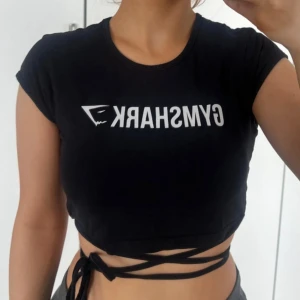 Gymshark <3 - Svart träningstopp från Gymshark med vit logga framtill. Toppen har korta ärmar och långa knytband som kan lindas runt midjan för en snygg detalj 🖤