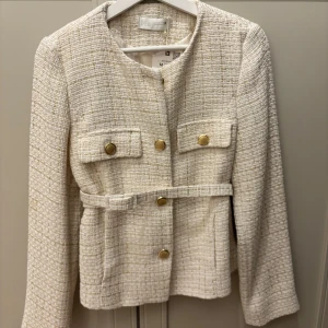 Off-whiite kavaj med guldknappar från Zara - Snygg kavaj från Zara med guldiga knappar och två fickor framtill. Jackan har ett vävt tyg med inslag av guldtråd och ett bälte i midjan för extra stilpoäng. Rund halsringning och långärmad modell, perfekt för att lyfta din outfit.