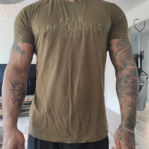 Olivgrön Peak Performance t-shirt - Snygg olivgrön t-shirt från Peak Performance med broderad logga framtill. Klassisk rund hals och korta ärmar. Skön passform och tillverkad i mjuk bomull som känns nice mot huden. Perfekt för dig som gillar stilrena och sportiga plagg.