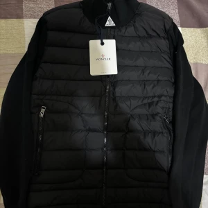 Svart dunjacka från Moncler - Svart dunjacka från Moncler med quiltad front och stickade ärmar. Jackan har dragkedja framtill och två sidofickor med dragkedja. Klassisk Moncler-logga på ärmen och ribbad krage. Perfekt för kyliga dagar med stil.