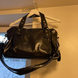 Svart axelväska i skinnimitation från SHEIN - Stilren svart axelväska med blanka metalldetaljer och bred axelrem. Väskan har två rejäla handtag med nitar och en stor öppning med dragkedja. Insidan är fodrad i tyg och har en innerficka med dragkedja. Perfekt för dig som vill ha plats för allt viktigt. Helt ny. Har fått två likadana, säljer den ena.