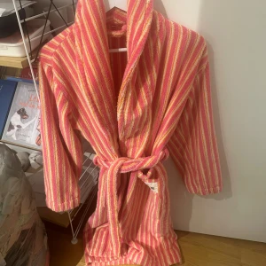 Randig frottémorgontock från Djerf Avenue - Säljer en mysig randig morgonrock från Djerf Avenue i färgerna rosa, orange och vit. Morgonrocken har lång ärm, bred krage och knyts med bälte i midjan. Perfekt för chill dagar hemma eller efter duschen. Märke syns på fickan. Finns några sömmar som har gått upp, se bild men inget som märks av