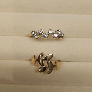 Två guldiga ringar med stenar och blad - Säljer två snygga guldiga ringar. Den ena ringen har glittrande stenar i rad och den andra har ett coolt bladmotiv. Perfekta accessoarer för att lyfta din stil och ge en trendig touch till dina outfits.