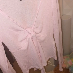 Ljusrosa omlottopp med knyt - Supersöt ljusrosa omlottopp med lång ärm och knyt framtill. Toppen har v-ringning och är tillverkad i mjukt bomullstyg. Perfekt för dig som gillar en feminin och enkel stil.