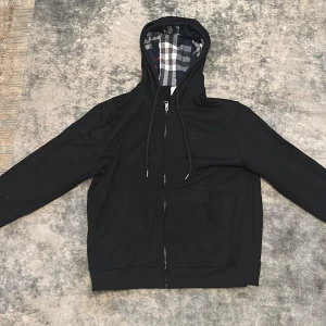 Svart hoodie med rutigt foder Burberry - Svart hoodie från Burberry med dragkedja och klassiskt rutigt foder i huvan. Tröjan har snörning i huvan, två fickor framtill och långa ärmar. Perfekt för en chill och stilren look.