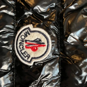 Svart pufferväst  från Moncler - Säljer en svart, glansig pufferväst  från Moncler i storlek L. Jackan har quiltad design med Moncler-logga på bröstet och coolt band med tryck på insidan. Perfekt för dig som gillar streetstyle och vill ha något som sticker ut.