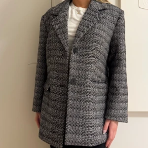 Svartvit rutig kavajjacka från Kaffe - Snygg svartvit kavajjacka från Kaffe med klassiskt rutig mönster i fiskbensstil. Jackan har två stora knappar framtill, breda slag och fickor med lock. Materialet är mjukt och känns som ullmix, perfekt för dig som gillar stilrena och tidlösa plagg.