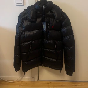 Svart pufferjacka från Polo Ralph Lauren - Snygg svart pufferjacka från Polo Ralph Lauren med röd logga på bröstet. Jackan har huva, flera dragkedjefickor och justerbara muddar med knapp. Materialet är glansigt och jackan är fodrad för extra värme. Perfekt för kalla dagar och har en modern, sportig vibe.Kom dm för frågor🧐, pris kan diskuteras😁. 