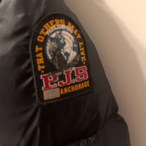 Svart pufferjacka Parajumpers XS - Snygg svart pufferjacka från Parajumpers, modell Sheen. Jackan har en glansig finish, huva, två stora fickor med lock och två snedställda dragkedjefickor. Märkespatch på ärmen och orange detalj vid kragen. Perfekt för kalla dagar.