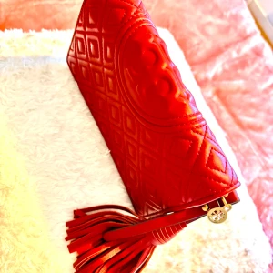 Röd quiltad axelväska Tory Burch - Snygg röd axelväska från Tory Burch i quiltat skinn med diamantmönster. Väskan har guldfärgad kedja, läderrem och en stor tofsdetalj. Logotypen är präglad på framsidan och insidan har magnetlås. Perfekt för dig som gillar statement-accessoarer.