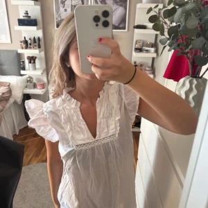 Vit kortklänning med spets från 157 - Supersöt vit kortklänning från 157 i storlek XS. Klänningen har volangdetaljer vid axlarna, v-ringad hals och dekorativ spets över bröstet. Tillverkad i mjuk bomull, perfekt för sommarens alla tillfällen. Luftig och lätt med romantiska vibbar.