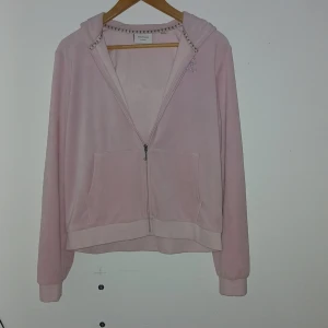 Rosa Juicy Couture hoodie - Säljer en ljusrosa hoodie från Juicy Couture i mjuk velour med strassdetaljer på bröstet och logga i strass på ryggen. Tröjan har huva, dragkedja, två fickor fram och långa armar. Perfekt för dig som gillar den klassiska Y2K-stilen!
