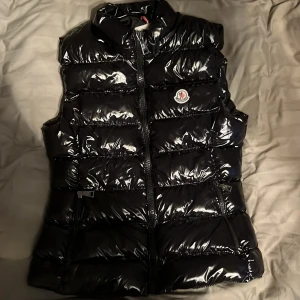 Svart dunväst från Moncler - Snygg svart dunväst från Moncler med glansig finish och quiltad design. Västen har dragkedja framtill, två sidofickor med dragkedja och klassisk Moncler-logga på bröstet. Perfekt för lager-på-lager under kyliga dagar.